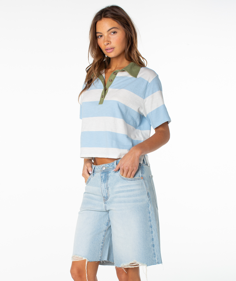 A woman stands facing forward against a plain white background wearing the Roxy Brookview Polo, a bold blue and white striped short-sleeve shirt with a relaxed fit, paired with light denim knee-length shorts.