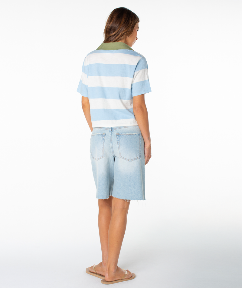A person stands facing away, wearing the Roxy Brookview Polo in relaxed fit with bold blue-and-white stripes, paired with light-wash denim shorts and beige sandals against a plain white background.