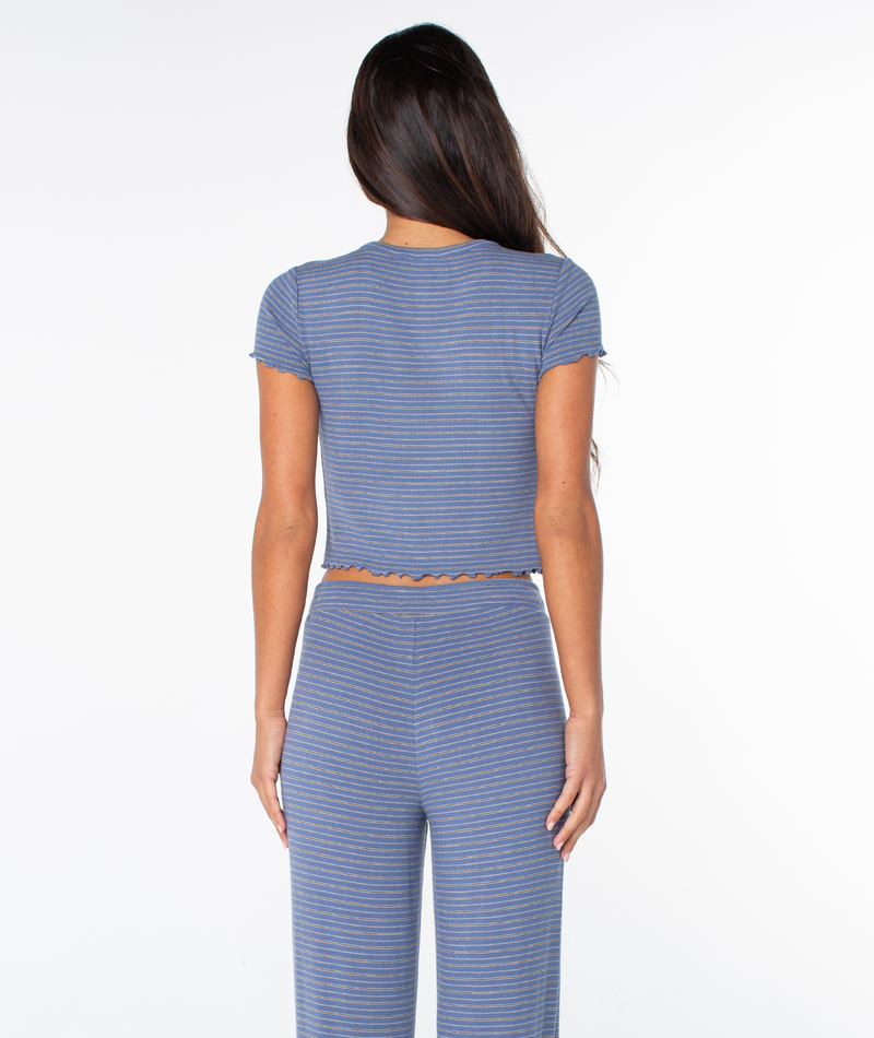 A person with long brown hair faces away, wearing the Poolside Crop Tee by Roxy—blue and gray striped, cropped fit—with matching pants against a white background.