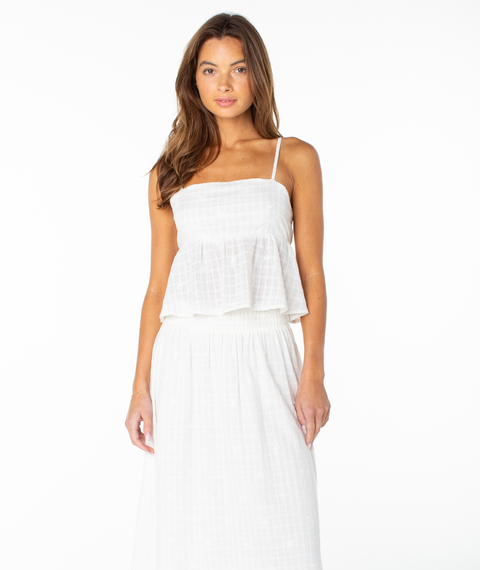 A woman wearing the Roxy Pierside Top in Coconut Milk Solid—an elegant, tank-style white sleeveless top with thin straps and a flowy peplum—paired with a matching white skirt and standing against a white background.
