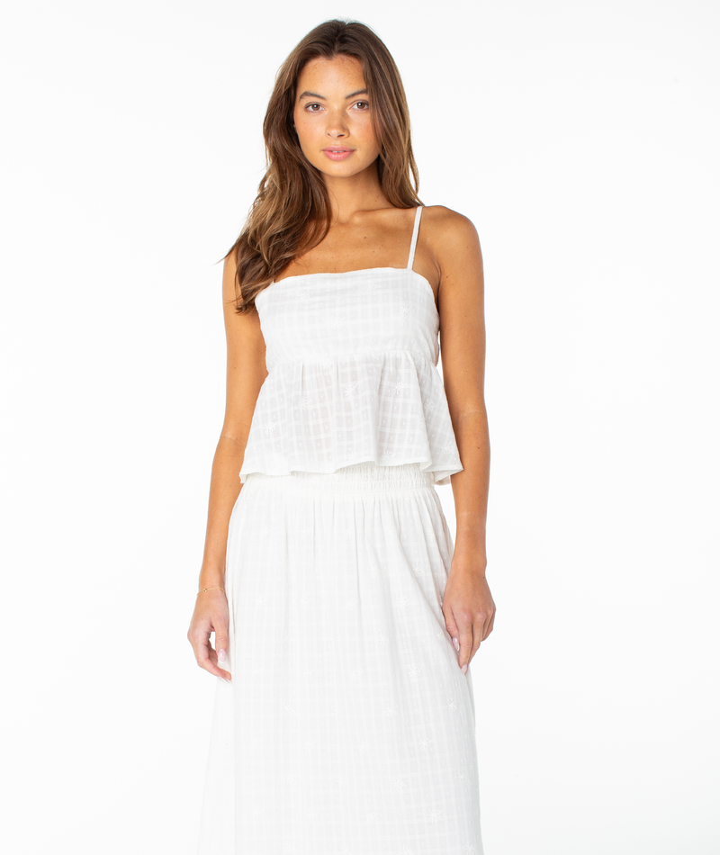 A woman wearing the Roxy Pierside Top in Coconut Milk Solid—an elegant, tank-style white sleeveless top with thin straps and a flowy peplum—paired with a matching white skirt and standing against a white background.
