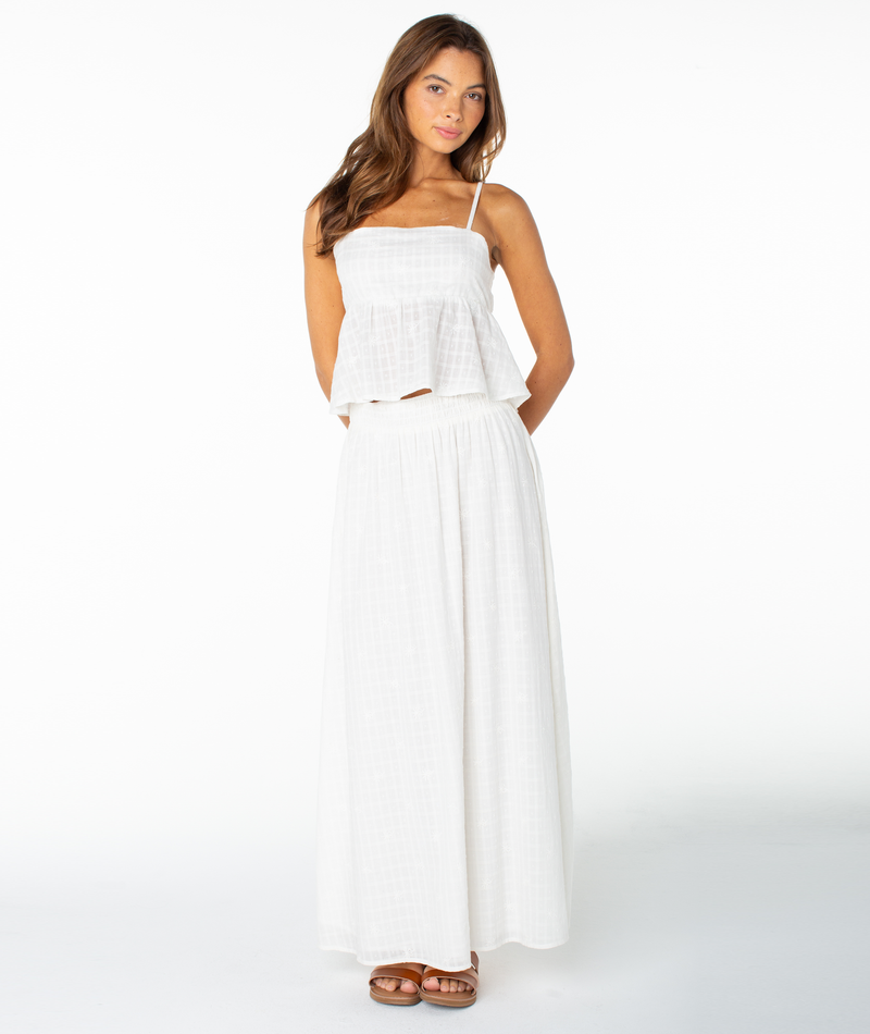 A woman wearing the Roxy Pierside Top—a flattering, white, sleeveless spaghetti strap top—paired with a long, flowy white skirt stands against a plain white background, looking slightly to the side with her hands behind her back.