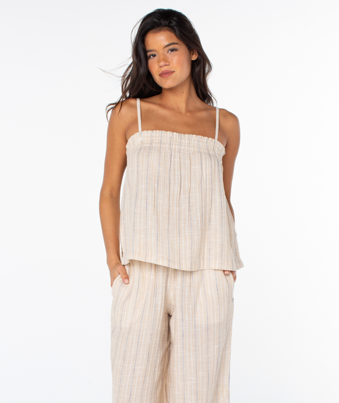 A woman stands against a white background wearing the Roxy Offshore Top in Doeskin Parchment Tbd Stripe—sleeveless and loose-fitting with thin straps. She pairs it with matching wide-leg pants, hands in pockets—ideal for a tropical getaway.