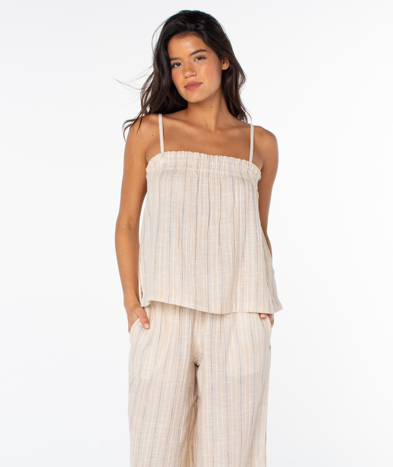 A woman stands against a white background wearing the Roxy Offshore Top in Doeskin Parchment Tbd Stripe—sleeveless and loose-fitting with thin straps. She pairs it with matching wide-leg pants, hands in pockets—ideal for a tropical getaway.