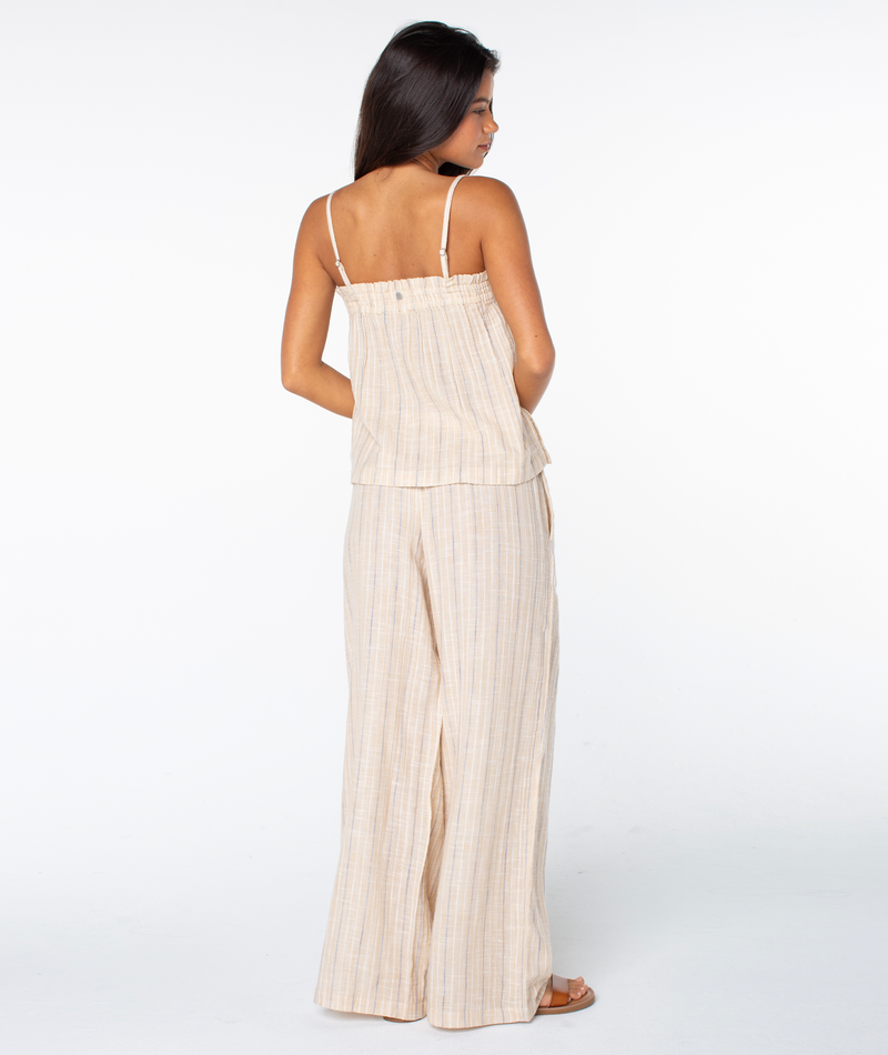 A woman faces away, wearing the Roxy Offshore Top—a sleeveless light beige plaid set with thin straps and matching wide-leg pants. With long dark hair and sandals, she’s styled for a tropical getaway against a white backdrop.