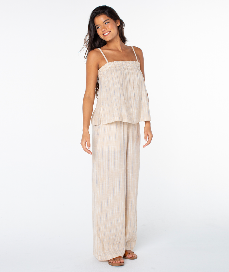 A woman with long dark hair smiles barefoot against a white background, wearing the Roxy Offshore Top with spaghetti straps and matching wide-leg pants—an ideal relaxed outfit for a tropical getaway.