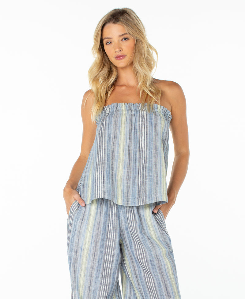 A woman with long blonde hair stands with her hands in her pockets against a white background, wearing the Roxy Offshore Top, which features a blue and gray striped strapless neckline, paired with matching wide-leg pants.