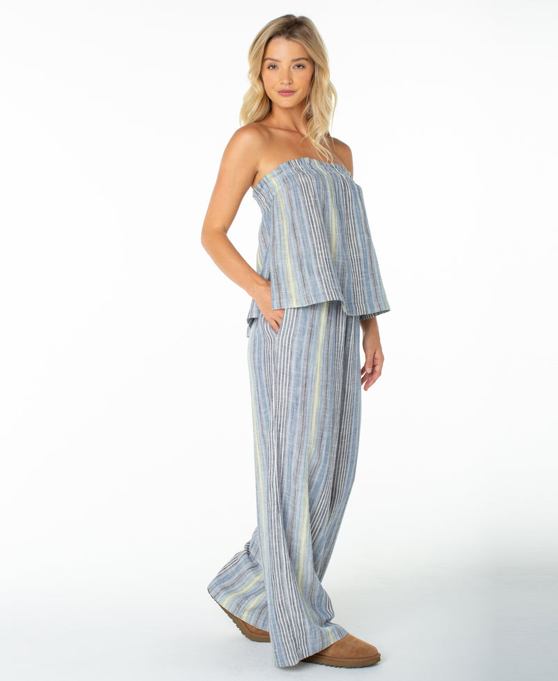 A woman is wearing the Roxy Offshore Top, a strapless, loose-fitting blue and gray striped top with a stylish neckline, paired with wide-leg pants and brown shoes, standing against a plain white background—perfect for a tropical vacation.