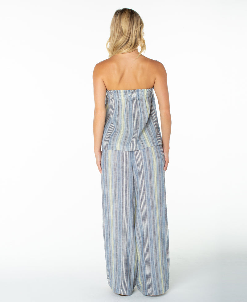 A woman with blonde hair stands facing away against a white background, wearing the Roxy Offshore Top—a strapless, loose-fitting top in blue and gray stripes—paired with matching wide-leg pants. This look is perfect for a tropical vacation.