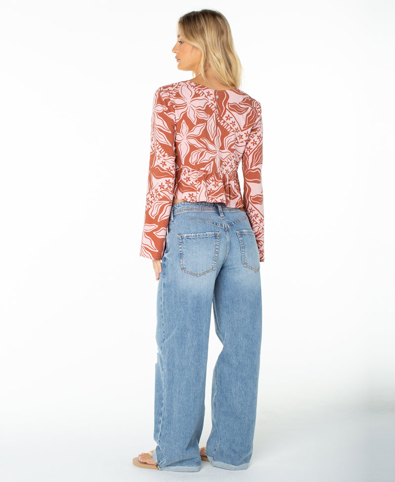 A woman stands with her back to the camera, wearing the Roxy Night Sky Top—a long-sleeve brown and white floral coastal shirt with a V neckline—paired with light blue wide-leg jeans and sandals, set against a plain white background.