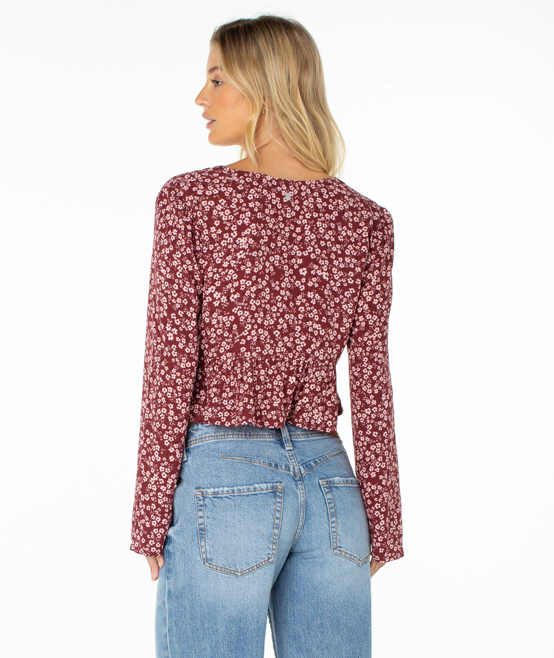 A woman with blonde hair, wearing the Roxy Night Sky Top—a maroon, long-sleeve floral blouse with a subtle V neckline—and light blue jeans stands facing away from the camera against a plain white background.