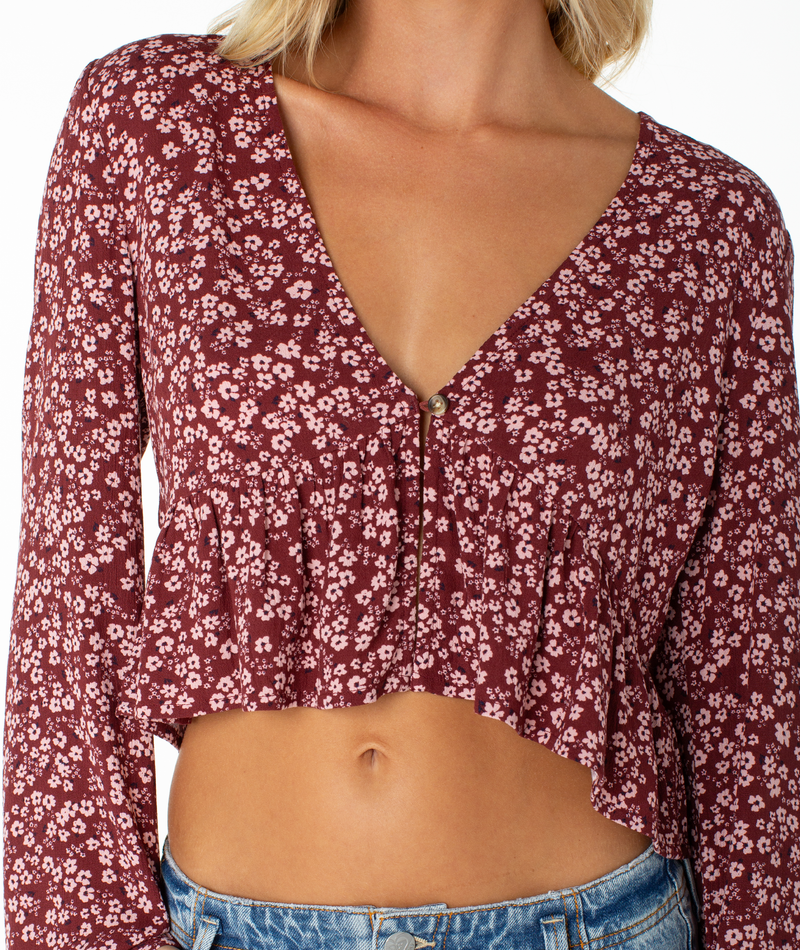 A person wearing the Roxy Night Sky Top—a maroon long-sleeve blouse with a deep V neckline, ruffled hem, small white floral print, and an elegant cropped drape—paired with blue jeans.