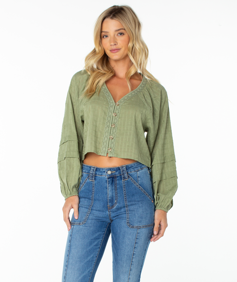 A woman with long blonde hair stands against a plain white background, wearing the Roxy Monterey Top in Oil Green Solid—a cropped button-front blouse with oversized sleeves—paired with high-waisted blue jeans.