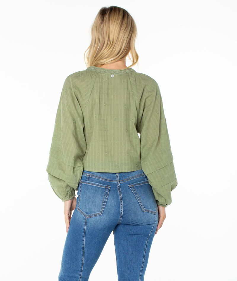 A woman with blonde hair, dressed in a Roxy Monterey Top in olive green featuring oversized sleeves and blue jeans, stands facing away from the camera against a white background.