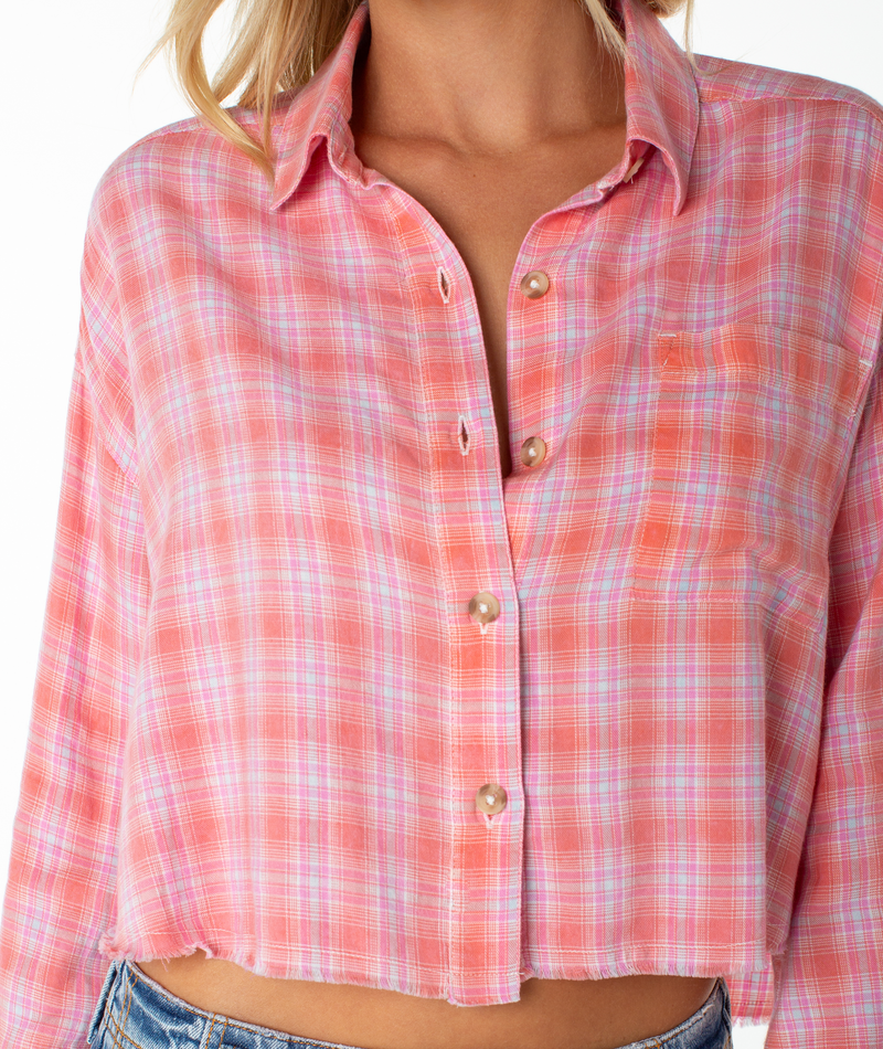 A person wears the Roxy Rockford Top, a pink and white plaid flannel button-up with frayed edges and a chest pocket, slightly open at the collar. The shirt is paired with blue denim jeans; image cropped at shoulders and waist.