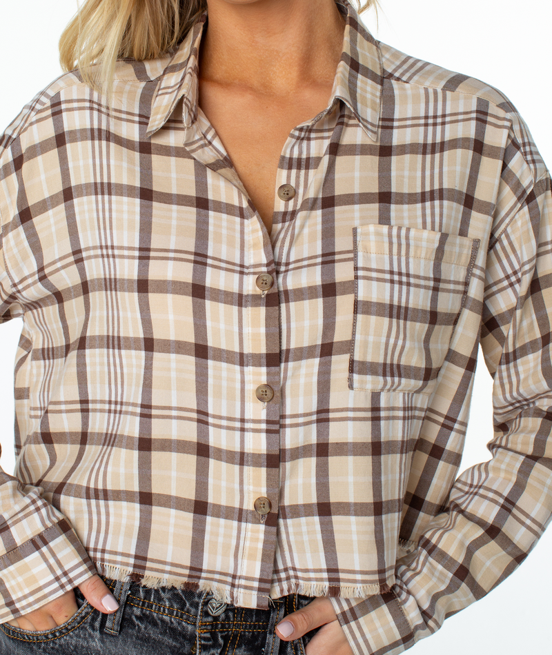 A person wears the Roxy Rockford Top—an oversized beige and brown plaid button-up with chest pocket and frayed hem—standing with one hand in a jean pocket; only their torso and lower face are visible.