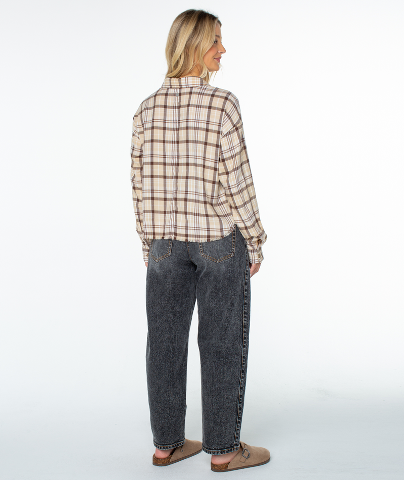 A woman stands facing away in the Roxy Rockford Top, an oversized beige plaid shirt, with loose dark gray jeans and tan slip-on shoes against a plain white background.