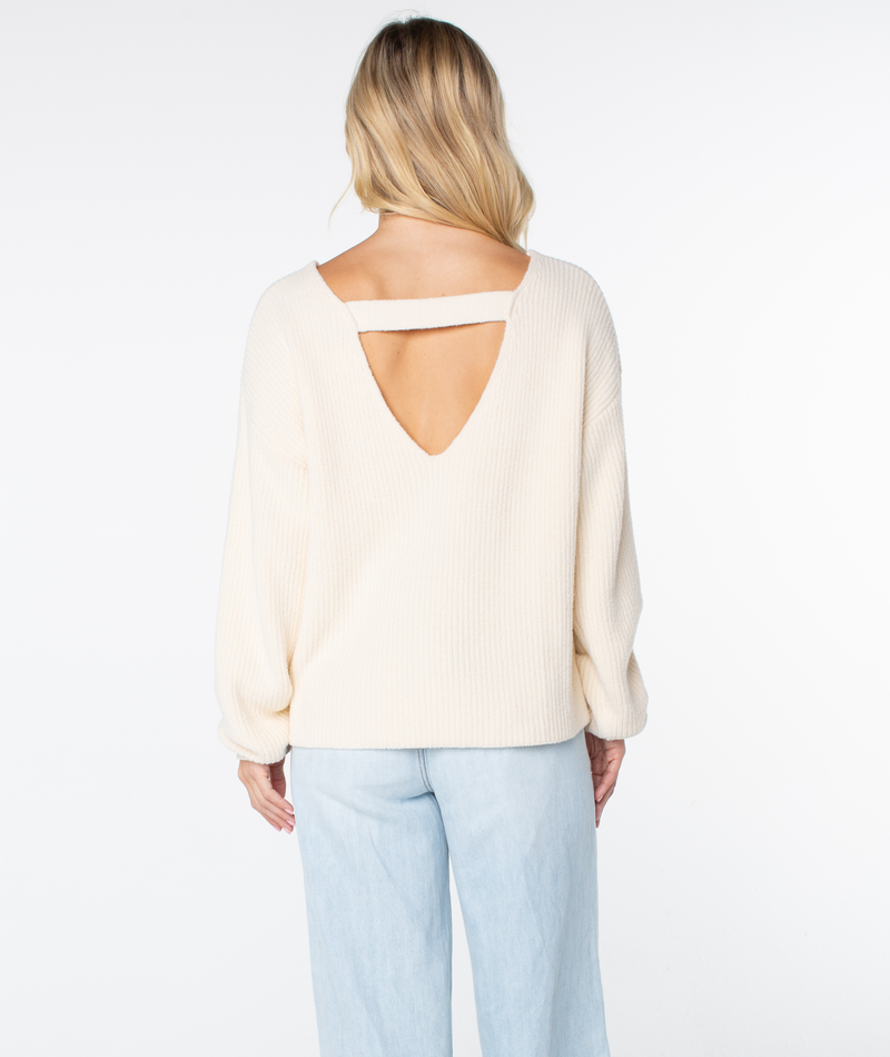 A woman faces away, wearing the Roxy Bombay 2 Sweater—a cream, long-sleeve top with an open back, V-cutout, and horizontal strap—paired with light blue jeans against a plain white background.