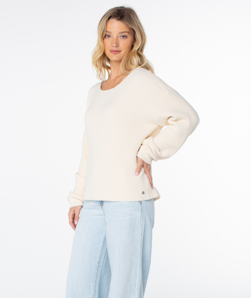 A woman with blonde hair wears the Roxy Bombay 2 Sweater and light blue jeans, standing against a plain white background, looking at the camera with a slight smile.