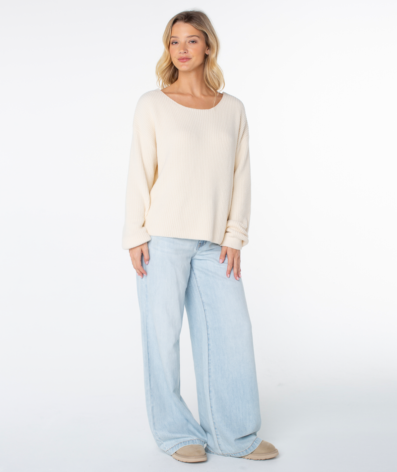 A woman with blonde hair stands against a white background, wearing the Roxy Bombay 2 Sweater in Parchment Solid, light blue wide-leg jeans, and beige shoes, looking relaxed with her arms by her sides.