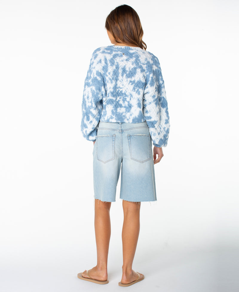A woman stands facing away against a plain white background, wearing the Roxy Sundaze Sweater—a blue and white dip-dye knitwear piece—paired with light-wash denim Bermuda shorts and tan slide sandals.