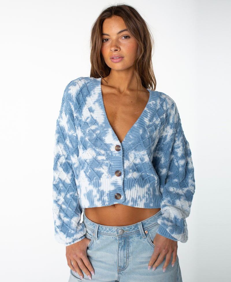 A woman wearing the Roxy Sundaze Sweater in Dutch Blue Tie Dye with a button front and light denim jeans stands against a plain white background, looking at the camera.