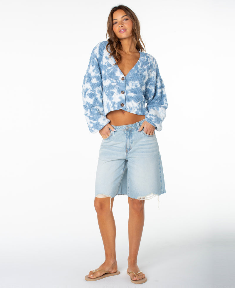 A woman stands against a white background, wearing the Roxy Sundaze Sweater—a blue dip-dye knitwear cropped sweater with a button front—paired with light denim knee-length shorts featuring a raw hem and beige flip-flops. Her wavy hair frames her face as she casually slips her hands into her pockets.