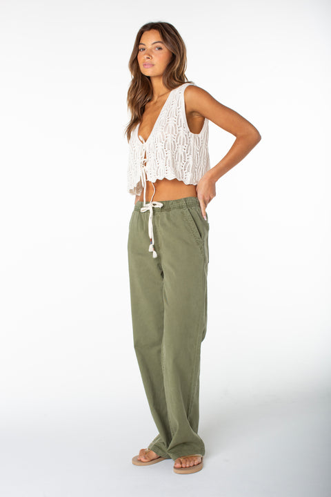 Woman in profile wears a white crochet sleeveless top and Roxy Sunset Dream Twill Pant in Four Leaf Clover Solid, hands in pockets, against a plain white background.