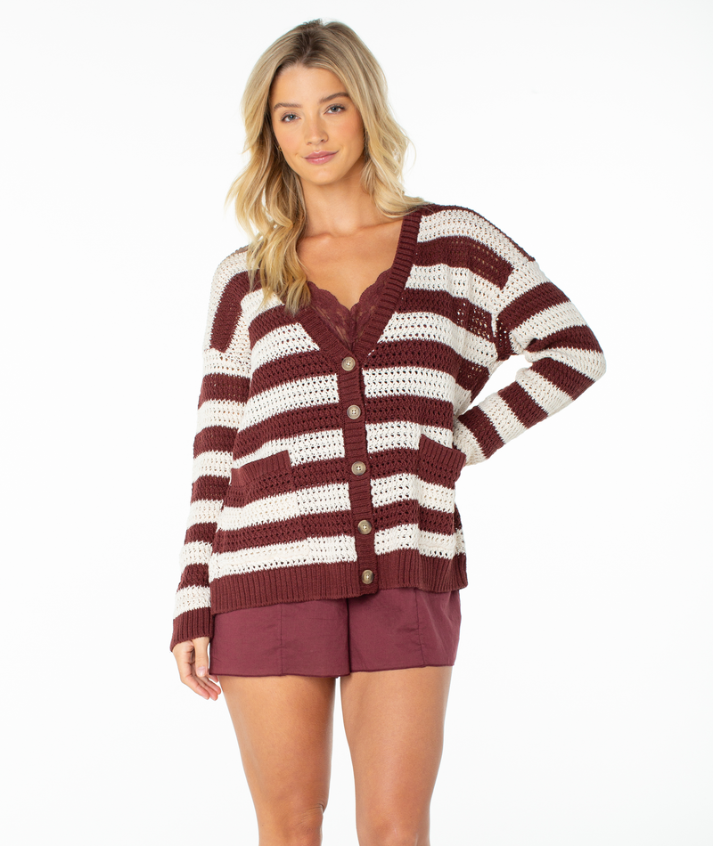 A woman stands against a white background, facing forward with one hand on her hip, wearing the Roxy Tamarindo Cardigan in Port Stripe over a maroon lace-trim top and matching shorts.