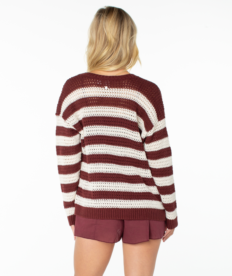 A person with long blonde hair is wearing the Roxy Tamarindo Cardigan—a maroon and white striped lightweight sweater—paired with maroon shorts, standing with their back to the camera against a white background.