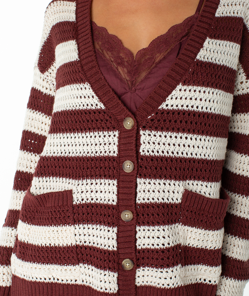 Wearing the Tamarindo Cardigan by Roxy—a maroon and white striped, lightweight sweater with buttons and two pockets—over a maroon lace-trimmed top.