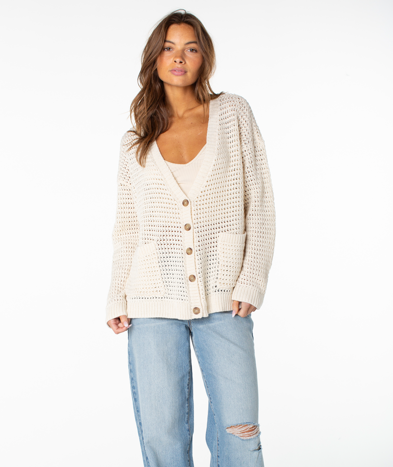 A woman wearing the Roxy Tamarindo Cardigan in Dew Stripe, featuring a chunky knit design with oversized buttons and front pockets, paired with light blue ripped jeans, stands against a plain white background.