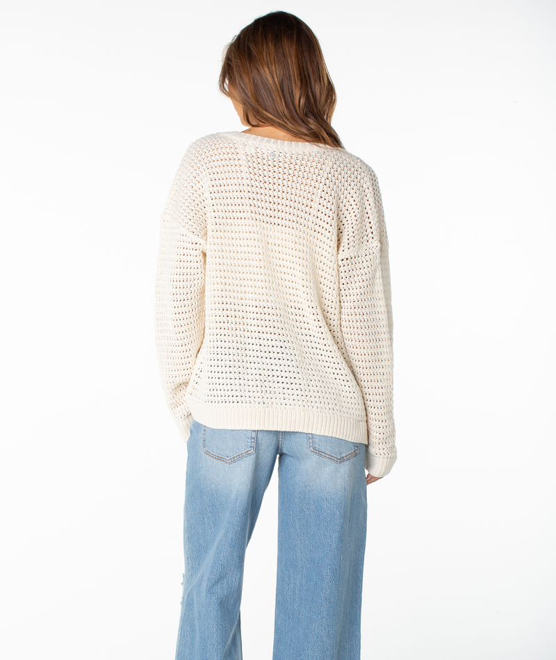 A woman stands facing away, wearing the Roxy Tamarindo Cardigan—a cream-colored, lightweight sweater with a relaxed V-neck—and light blue jeans. Her brown hair is down, and the background is plain white.
