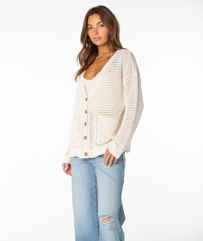 A woman with long brown hair stands against a white background, looking toward the camera while wearing the Roxy Tamarindo Cardigan—a cream knitted style featuring oversized buttons—layered over a matching top and paired with light blue ripped jeans.