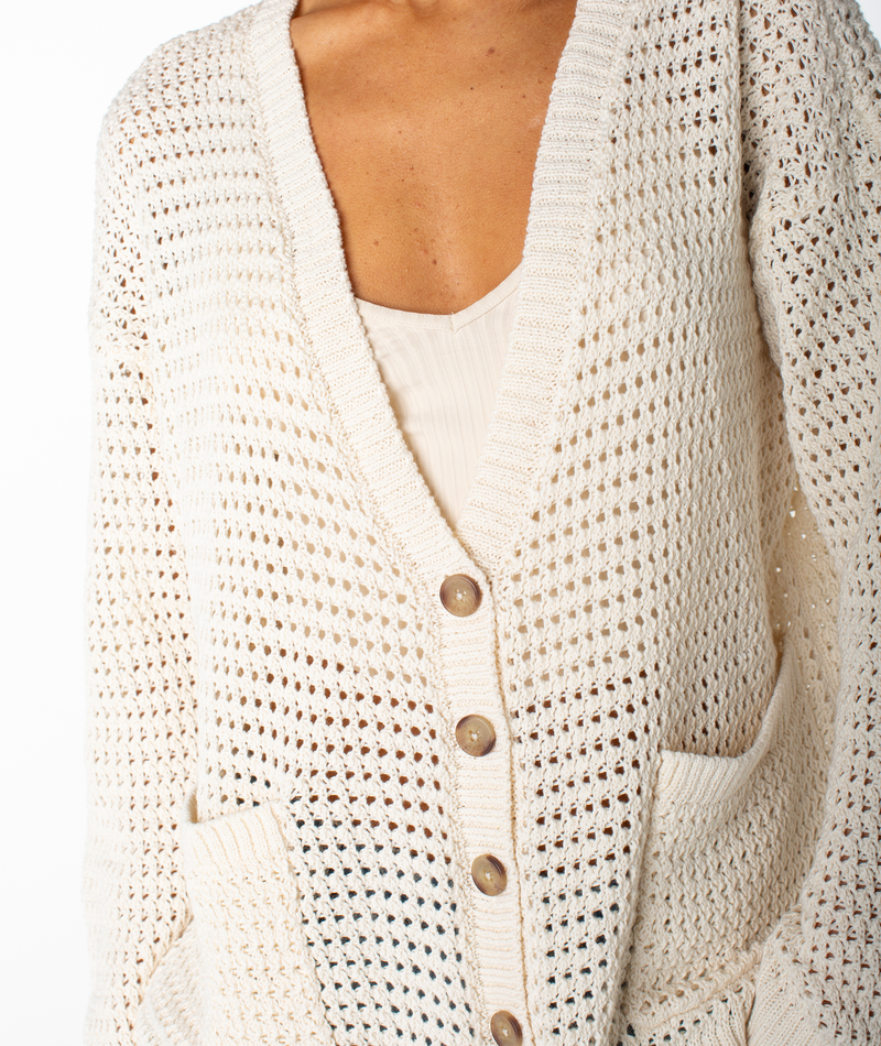 A person wearing the Roxy Tamarindo Cardigan, a cream-colored, open-knit style featuring oversized buttons and two front pockets, layered over a light top.