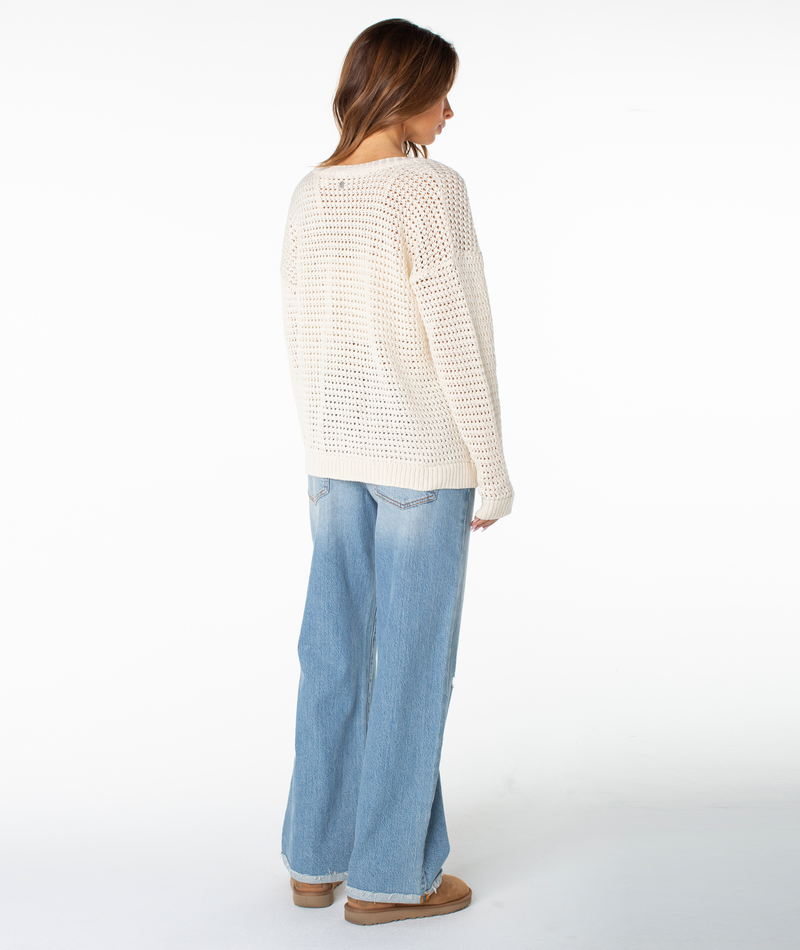 A woman with light brown hair stands facing away, wearing the Roxy Tamarindo Cardigan—a lightweight cream-colored knit sweater with oversized buttons—paired with light blue wide-leg jeans and tan shoes against a plain white background.