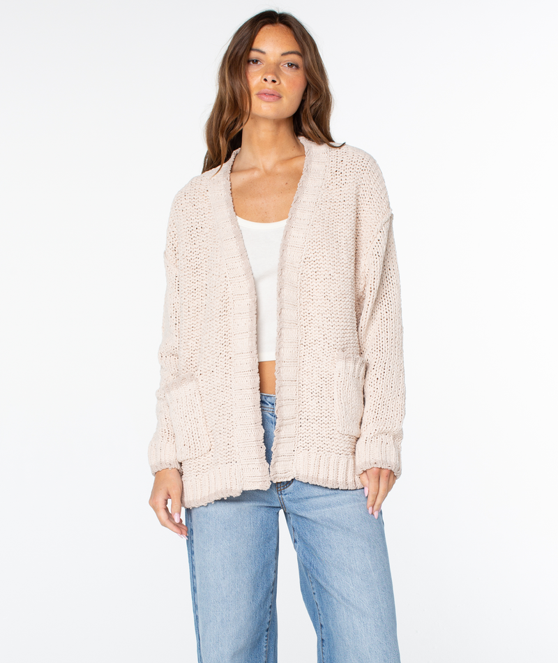 A woman wearing a Parchment 2 Solid Roxy Cozy Cruiser cardigan over a white tank top and blue jeans.