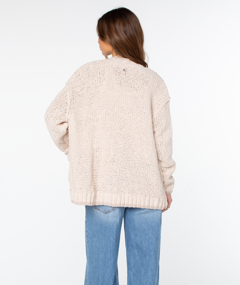 Viewed from behind, a person wears a cream-colored, chunky-knit Roxy Cozy Cruiser cardigan paired with blue jeans.