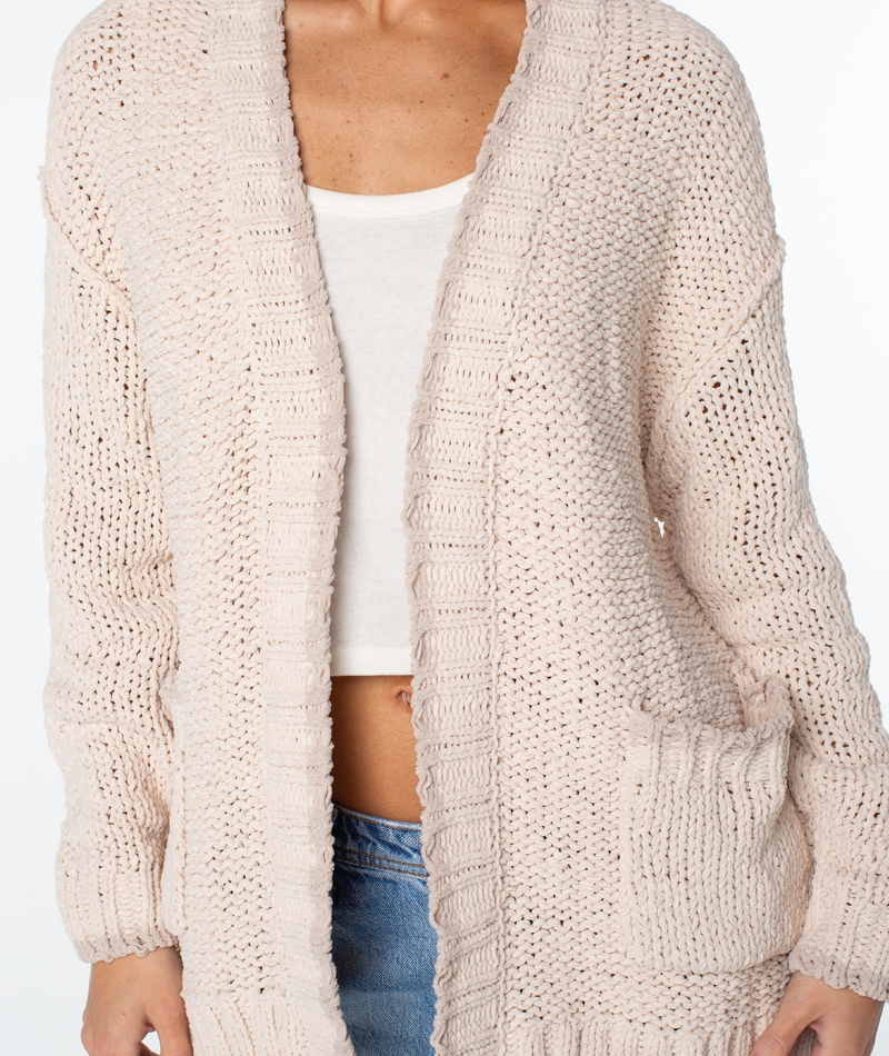 A person wearing an open, light-colored, chunky knit Roxy Cozy Cruiser cardigan over a white top and blue jeans.