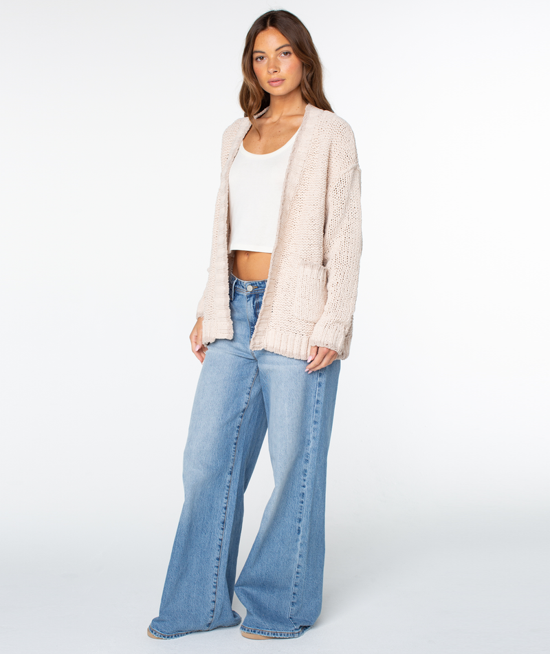 A person wearing a beige Roxy Cozy Cruiser cotton blend cardigan over a white tank top and wide-leg blue jeans.