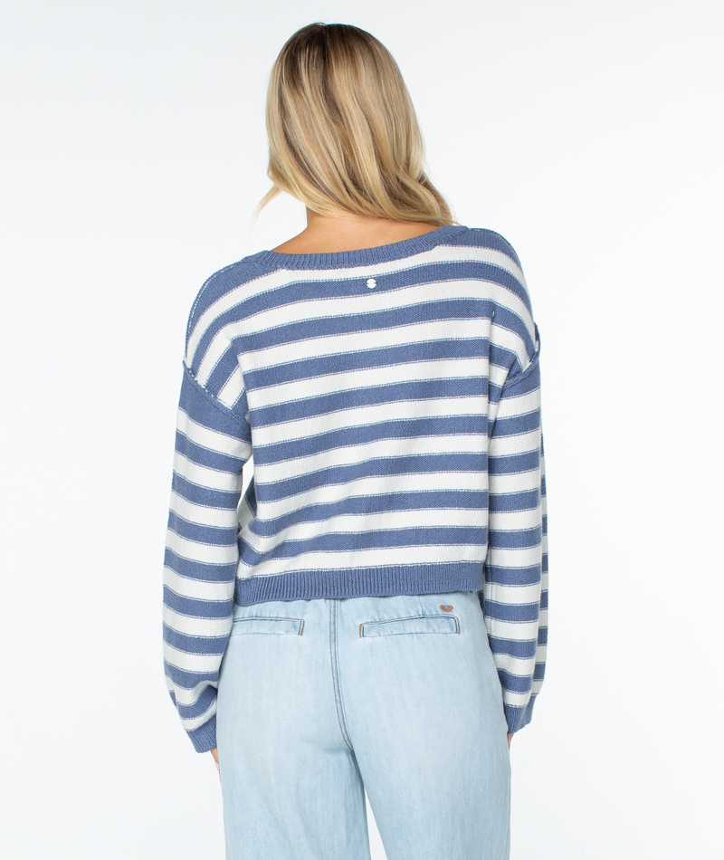 A woman with long blonde hair and light blue jeans wears the Roxy Swell Season Stripe Sweater, featuring blue and white stripes, as she faces away from the camera against a plain white background.