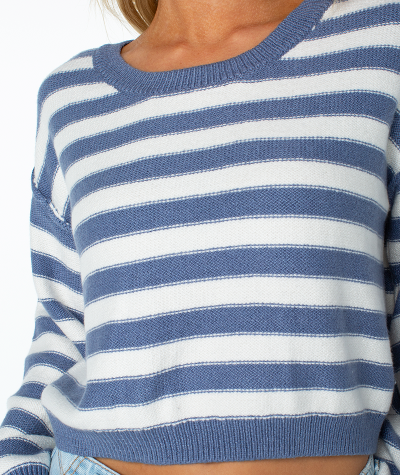 A person wears the Roxy Swell Season Stripe Sweater in blue and white stripes, cropped at the waist for a relaxed fit, paired with light denim jeans. The image highlights the cozy upper torso.