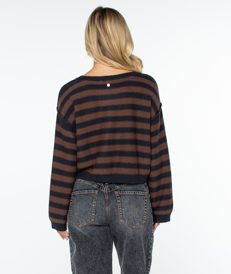 A person with long blonde hair, wearing the Roxy Swell Season Stripe Sweater in brown and black stripes and faded black jeans, stands facing away from the camera against a plain white background.