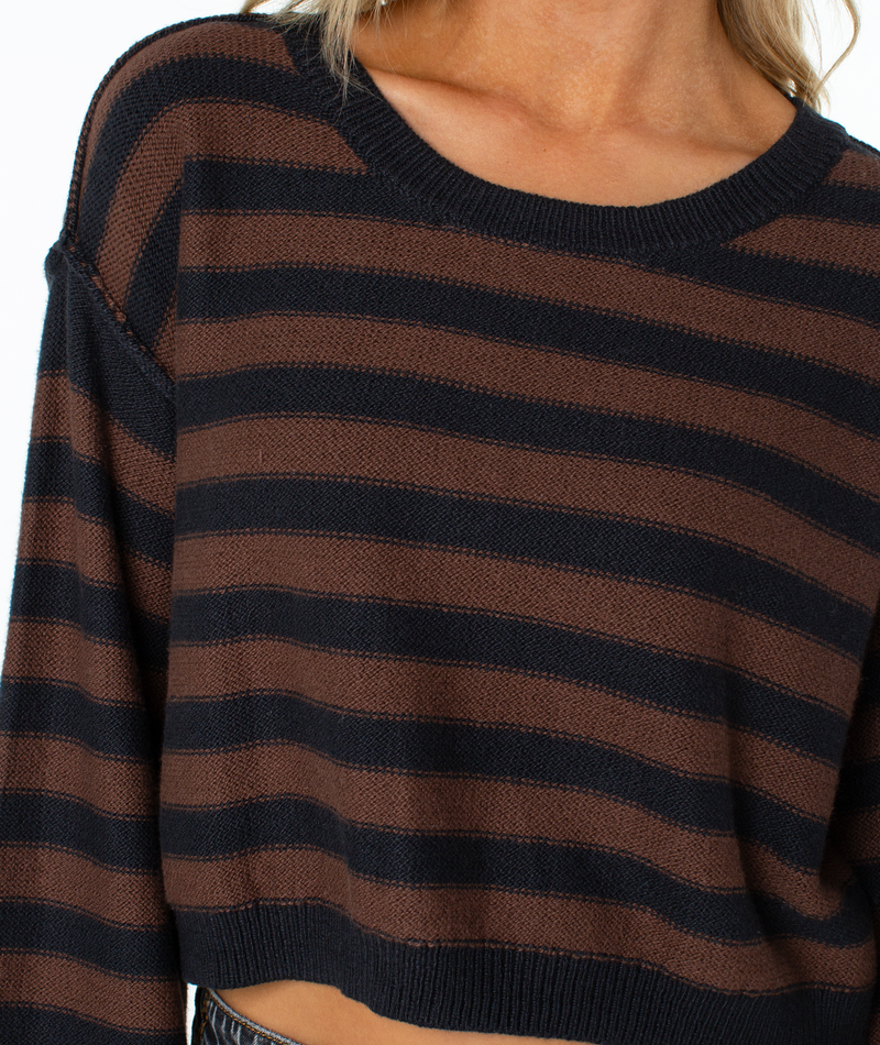 A person wears the Roxy Swell Season Stripe Sweater, a cozy brown and black striped, long-sleeve cropped sweater with a relaxed fit. Only the torso and part of the neck are visible.