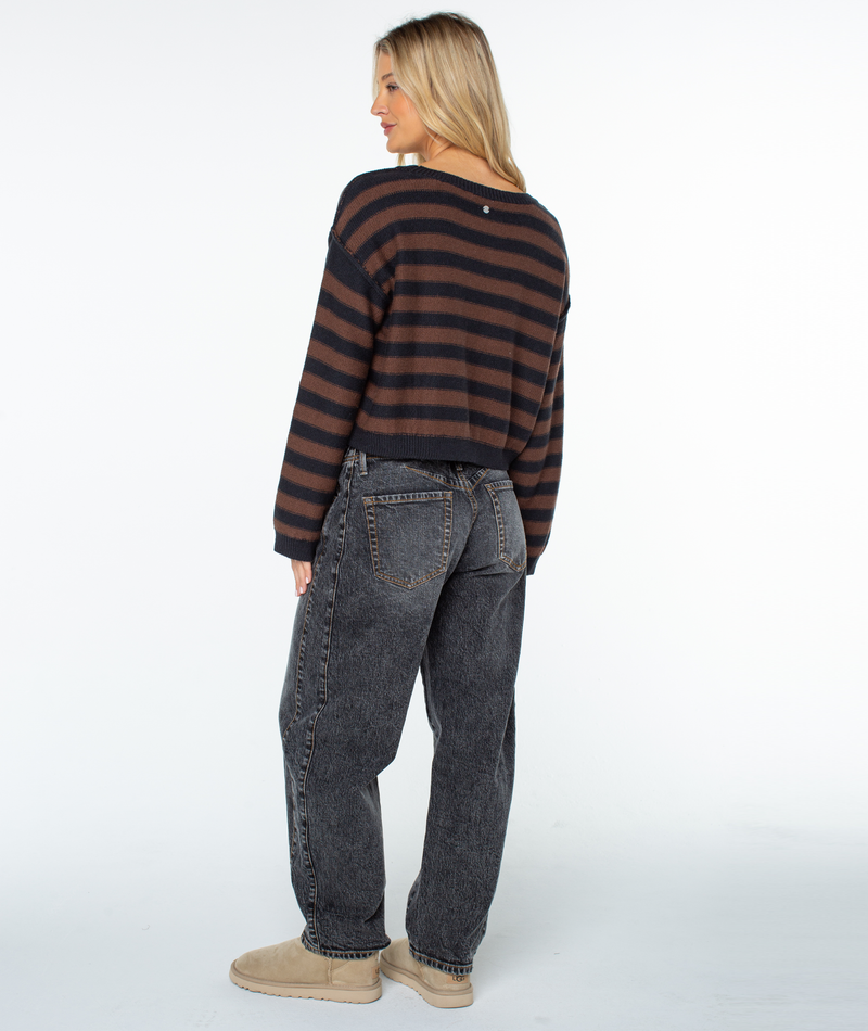 A person with long blonde hair wears the Roxy Swell Season Stripe Sweater with black and brown stripes, loose dark jeans, and beige shoes, standing and looking over their shoulder against a white background.