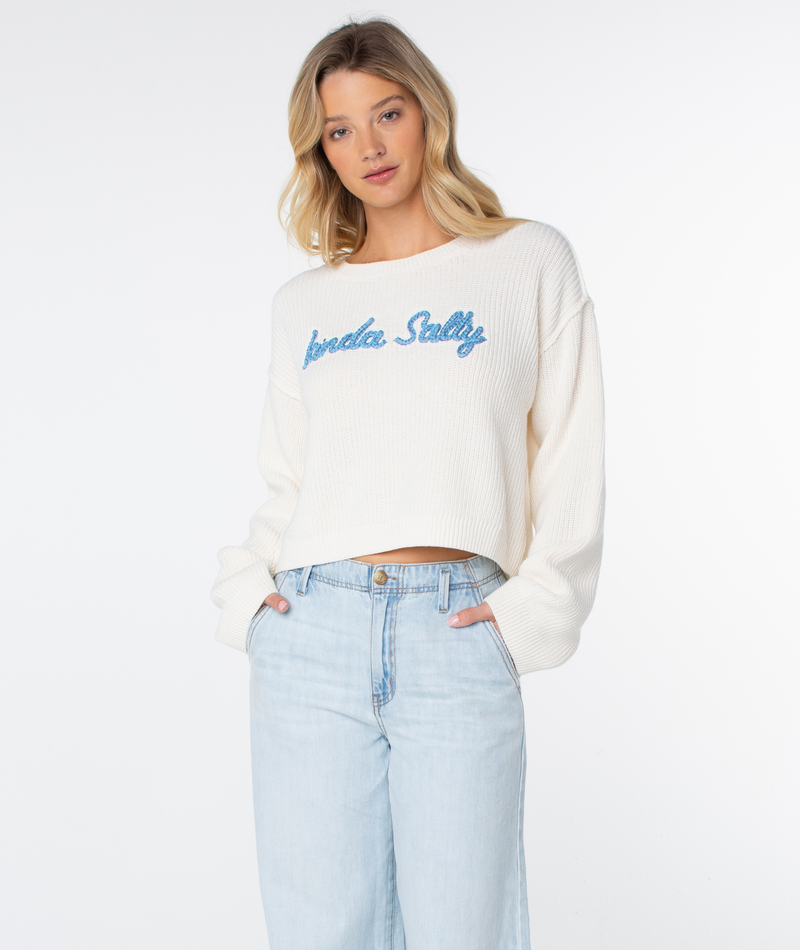 Woman with long blonde hair wears the Roxy Swell Season Sweater in Coconut Milk Kinda Salty and light-wash jeans, standing against a plain white background with hands in her pockets.