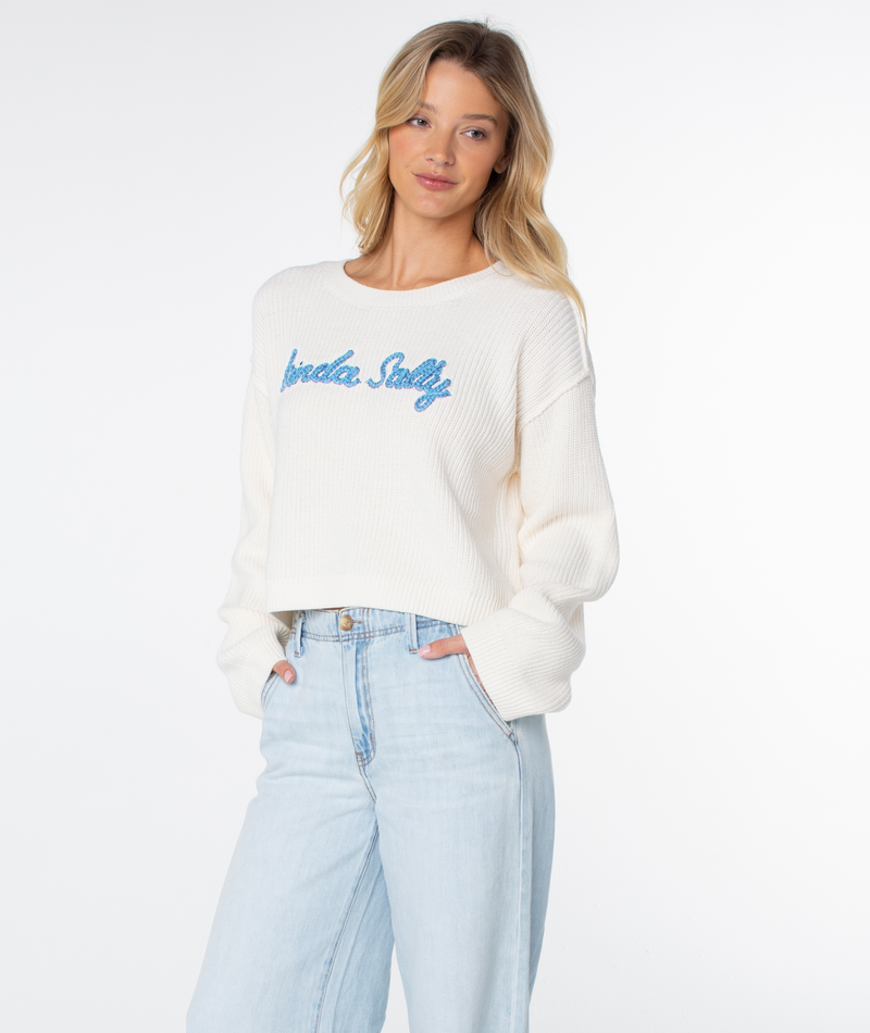 A person models the Roxy Swell Season Sweater in cream with blue text, paired with light blue jeans, standing against a white background and smiling gently at the camera.
