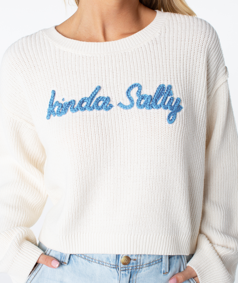 A person wears a cream-colored Swell Season Sweater by Roxy with kinda salty in blue textured letters, paired with light blue high-waisted jeans.