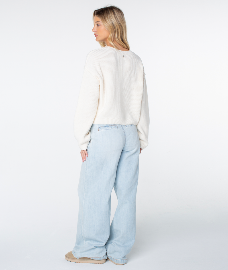 A woman with long blonde hair stands facing away, wearing the Roxy Swell Season Sweater, light blue wide-leg jeans, and beige shoes against a plain white background.