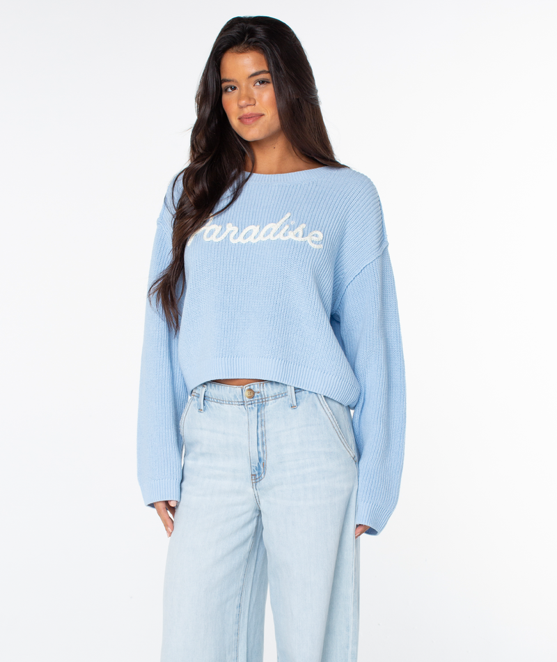 Woman with long brown hair wears a light blue Swell Season Sweater by Roxy with paradise text and light blue jeans, standing against a white background.